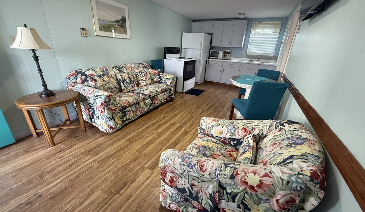 A small living area with wood flooring, floral-patterned sofa and armchair, round table with two chairs, and a compact kitchen with white appliances and light blue cabinets.