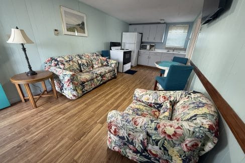 A small living area with wood flooring, floral-patterned sofa and armchair, round table with two chairs, and a compact kitchen with white appliances and light blue cabinets.