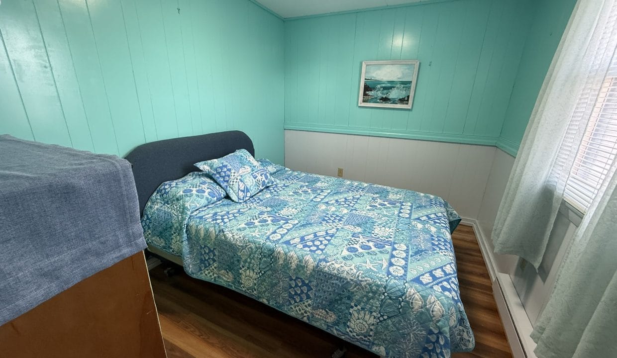 A small bedroom with turquoise walls, a bed covered in blue patterned bedding, a wooden floor, a window with light curtains, and a framed picture on the wall.
