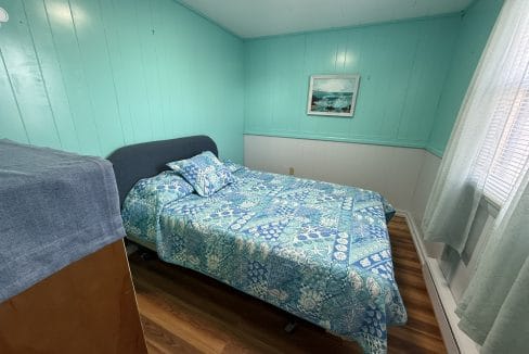 A small bedroom with turquoise walls, a bed covered in blue patterned bedding, a wooden floor, a window with light curtains, and a framed picture on the wall.