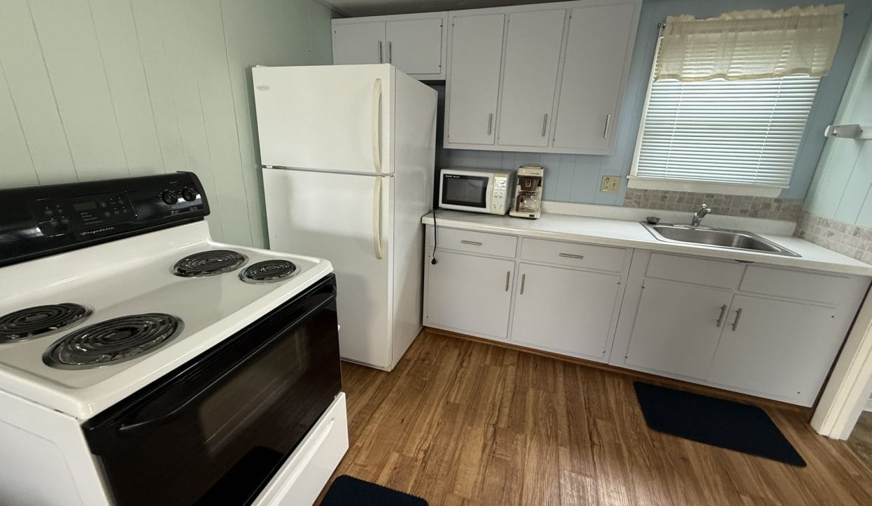 Small kitchen with a white stove, refrigerator, microwave, coffee maker, sink, and light blue cabinets, featuring wood flooring and a window with a sheer curtain.