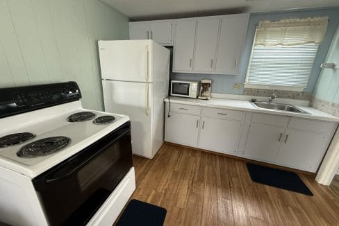 Small kitchen with a white stove, refrigerator, microwave, coffee maker, sink, and light blue cabinets, featuring wood flooring and a window with a sheer curtain.