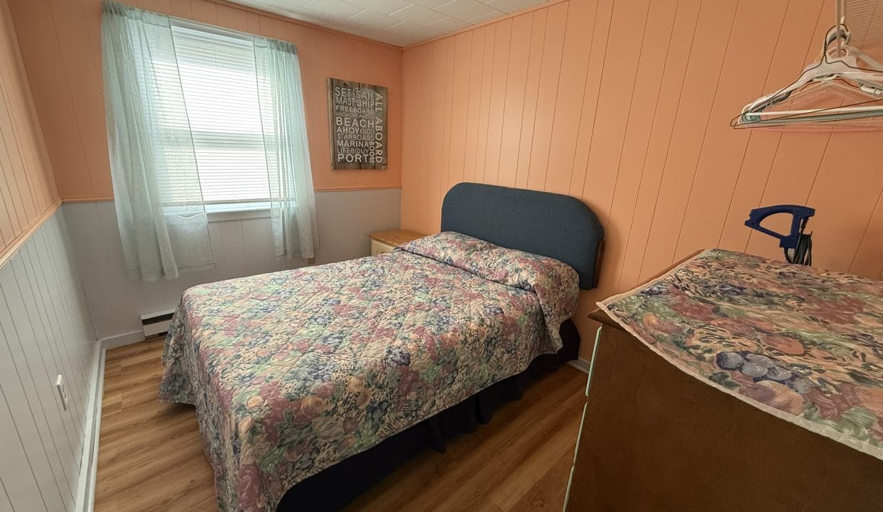 Small bedroom with peach-colored walls, a bed with floral bedding, a dresser with a matching cover, a window with light curtains, and a wall art piece.