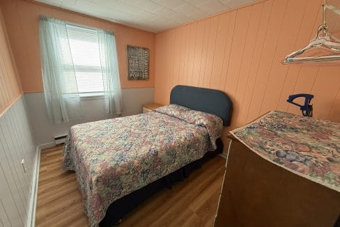 Small bedroom with peach-colored walls, a bed with floral bedding, a dresser with a matching cover, a window with light curtains, and a wall art piece.