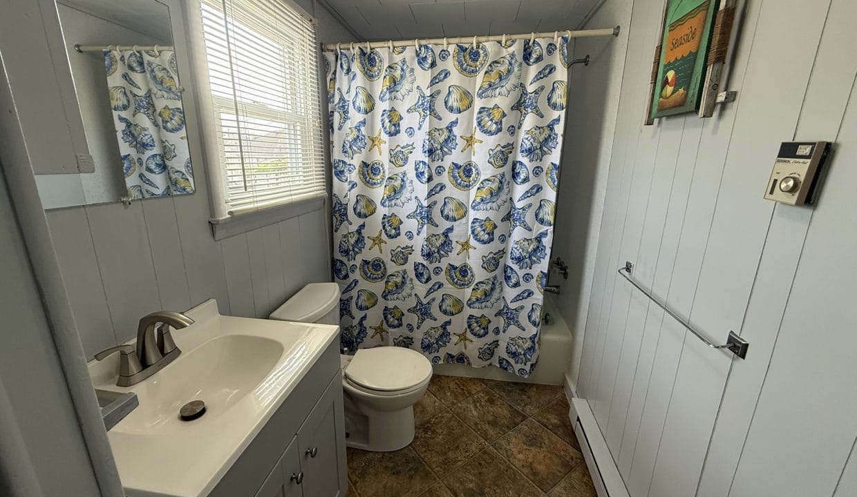 Small bathroom with light blue walls, a white sink and toilet, a colorful shower curtain with floral patterns, window with blinds, and a framed picture on the wall.