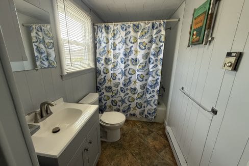 Small bathroom with light blue walls, a white sink and toilet, a colorful shower curtain with floral patterns, window with blinds, and a framed picture on the wall.