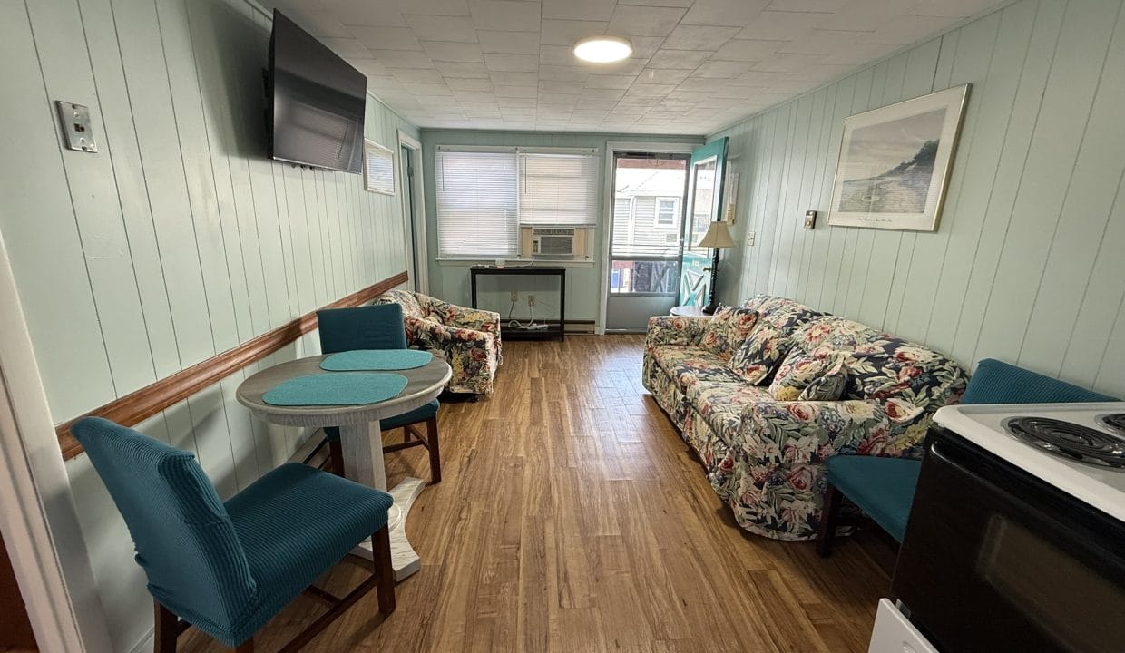 Small living space with wood flooring, floral sofa, teal chairs, wall-mounted TV, round table, and window air conditioner; walls are painted light green.