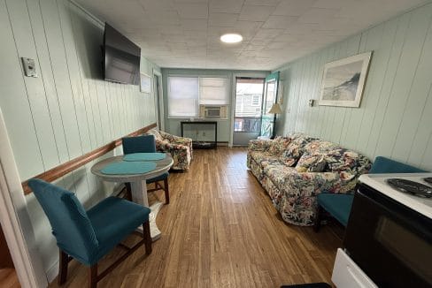 Small living space with wood flooring, floral sofa, teal chairs, wall-mounted TV, round table, and window air conditioner; walls are painted light green.
