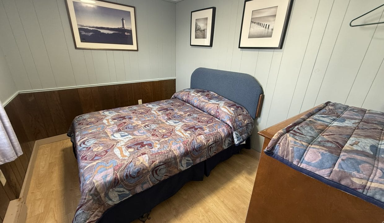 A small bedroom with a double bed, patterned bedspread, wooden dresser, framed photos on pale blue walls, and a wood floor.
