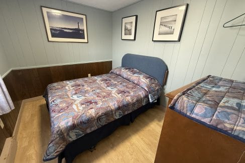 A small bedroom with a double bed, patterned bedspread, wooden dresser, framed photos on pale blue walls, and a wood floor.