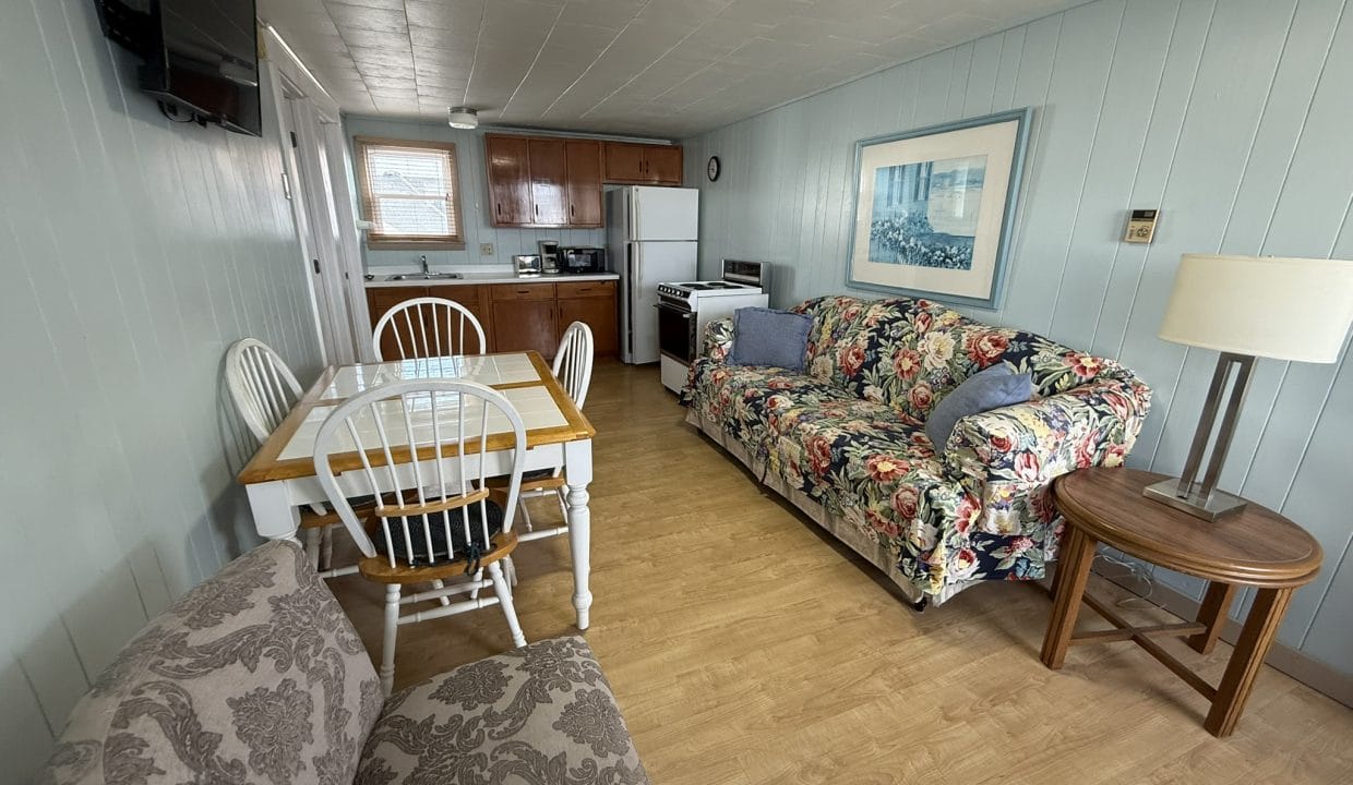 Small kitchen and living area with wood cabinets, dining table, patterned couch, armchairs, a side table, lamp, and wall-mounted TV. Light wood flooring and pastel blue paneled walls.