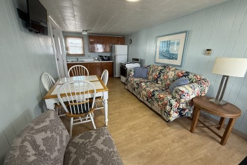 Small kitchen and living area with wood cabinets, dining table, patterned couch, armchairs, a side table, lamp, and wall-mounted TV. Light wood flooring and pastel blue paneled walls.