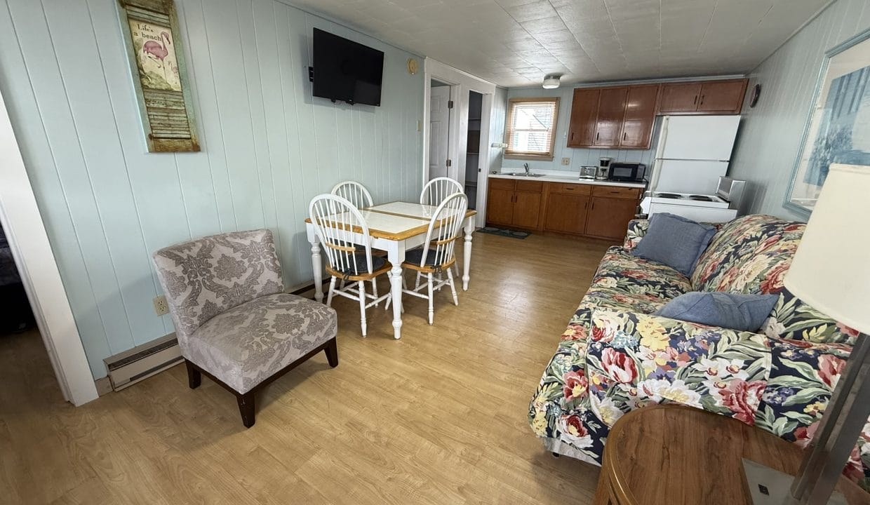 Small kitchen and dining area with wood floors, pale blue walls, a floral sofa, patterned armchair, dining table with four chairs, and basic kitchen appliances.