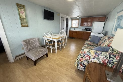 Small kitchen and dining area with wood floors, pale blue walls, a floral sofa, patterned armchair, dining table with four chairs, and basic kitchen appliances.