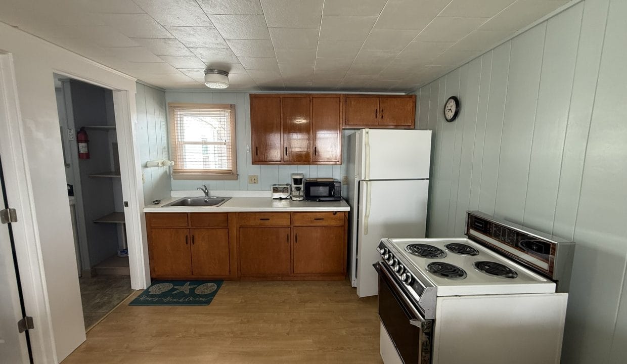 A small kitchen with wooden cabinets, white appliances including a stove and fridge, a sink under a window, and a clock on a light blue wall.