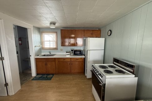 A small kitchen with wooden cabinets, white appliances including a stove and fridge, a sink under a window, and a clock on a light blue wall.