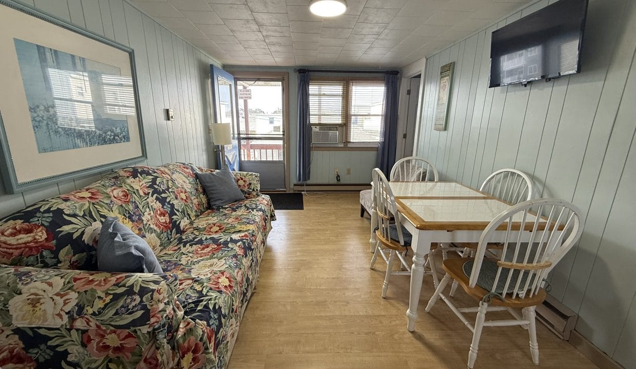 A small living and dining area with a floral sofa, a dining table with four chairs, wall-mounted TV, large window, and light wood flooring.