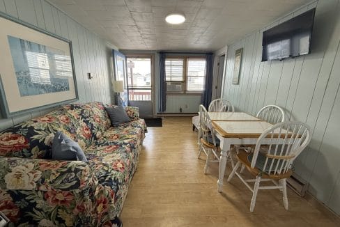 A small living and dining area with a floral sofa, a dining table with four chairs, wall-mounted TV, large window, and light wood flooring.