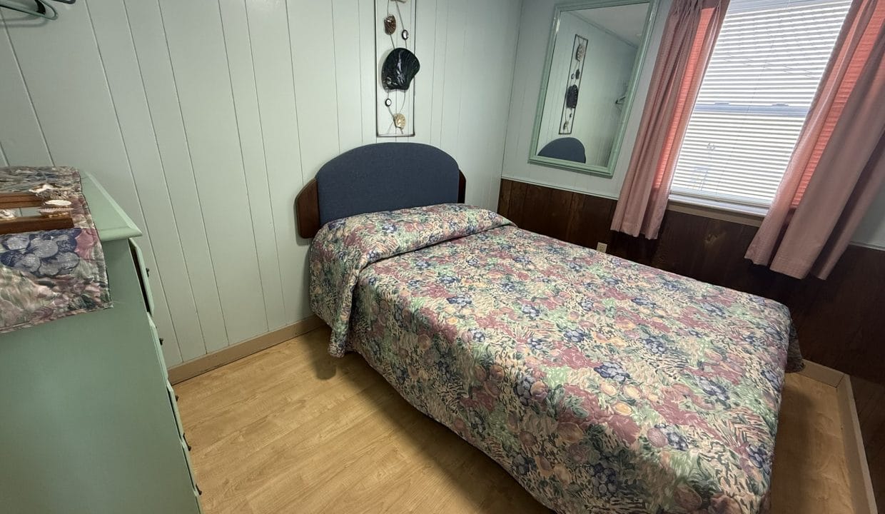 Small bedroom with wood panel walls, a bed with a floral bedspread, a dresser, a mirror, a window with pink curtains, and a wall decoration above the bed.