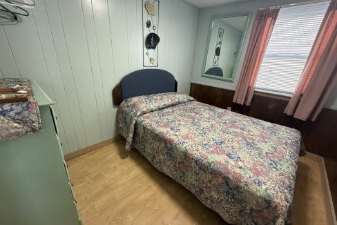 Small bedroom with wood panel walls, a bed with a floral bedspread, a dresser, a mirror, a window with pink curtains, and a wall decoration above the bed.