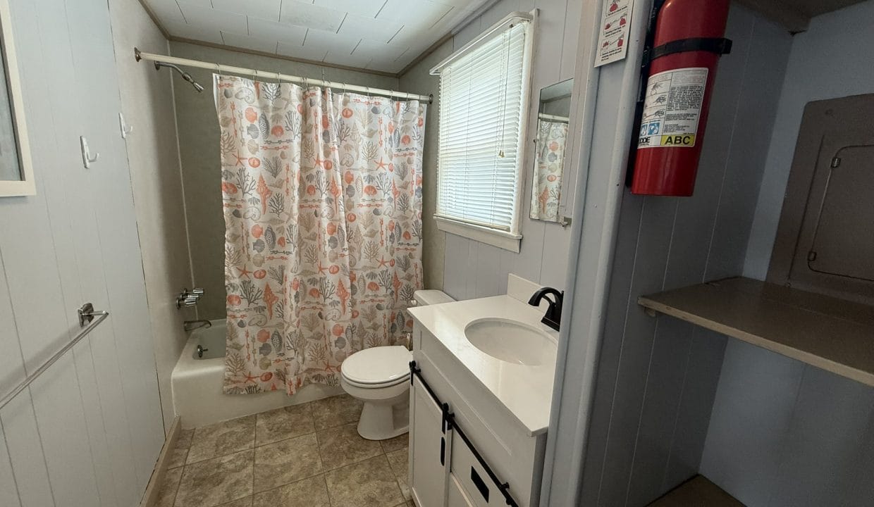 Small bathroom with a bathtub and shower curtain, toilet, white vanity with sink, wall mirror, window with blinds, open shelves, and a fire extinguisher mounted on the wall.