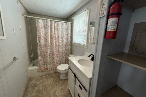 Small bathroom with a bathtub and shower curtain, toilet, white vanity with sink, wall mirror, window with blinds, open shelves, and a fire extinguisher mounted on the wall.