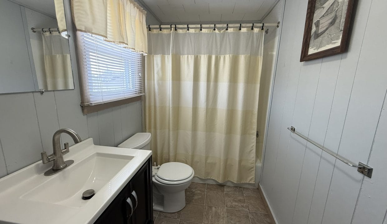 A small bathroom with a sink, mirror, toilet, towel rack, window with curtains, and a shower with a striped curtain.