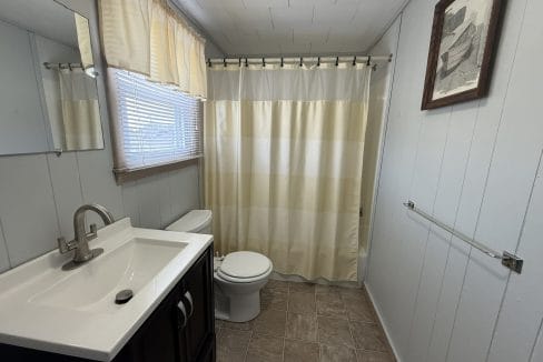A small bathroom with a sink, mirror, toilet, towel rack, window with curtains, and a shower with a striped curtain.