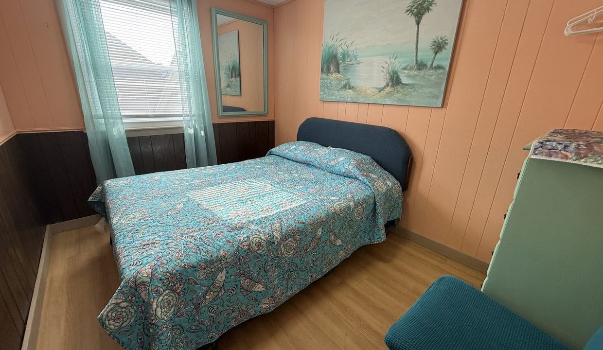 A bedroom with a double bed, blue and paisley bedding, teal chair, dresser, large beach painting, and window with sheer teal curtains.