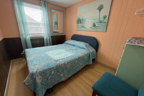 A bedroom with a double bed, blue and paisley bedding, teal chair, dresser, large beach painting, and window with sheer teal curtains.