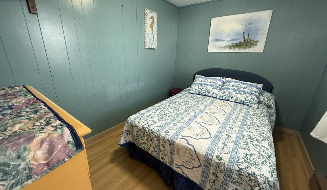 A small bedroom with teal wood-paneled walls, a bed with blue and white bedding, a wooden dresser, and two ocean-themed artworks on the walls.