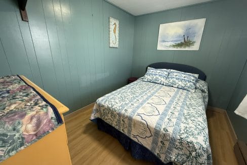 A small bedroom with teal wood-paneled walls, a bed with blue and white bedding, a wooden dresser, and two ocean-themed artworks on the walls.