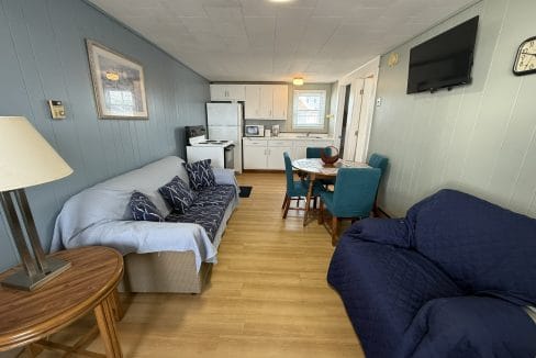 Small living and dining area with a sofa, armchair, dining table, and chairs; kitchen with white cabinets and appliances in the background. TV mounted on wall and wooden floors throughout.