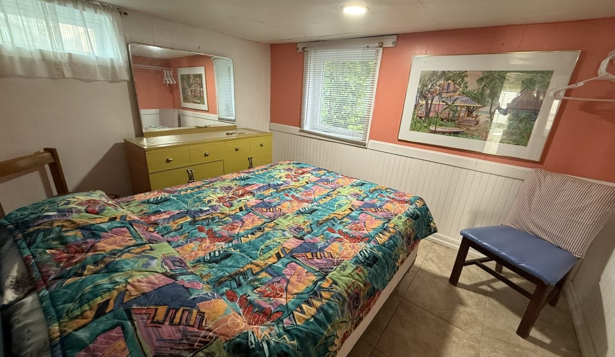 Small bedroom with a colorful quilt on the bed, a yellow dresser with mirror, a blue chair, a window with blinds, and a framed artwork on the salmon-colored wall.