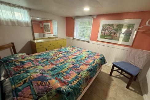 Small bedroom with a colorful quilt on the bed, a yellow dresser with mirror, a blue chair, a window with blinds, and a framed artwork on the salmon-colored wall.