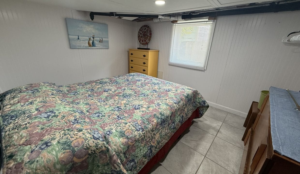 Small bedroom with a floral bedspread, a wooden dresser, a window with blinds, wall art, and tiled floor. The ceiling is low with exposed pipes.