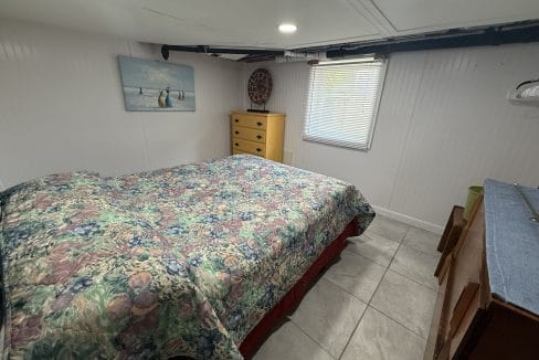 Small bedroom with a floral bedspread, a wooden dresser, a window with blinds, wall art, and tiled floor. The ceiling is low with exposed pipes.