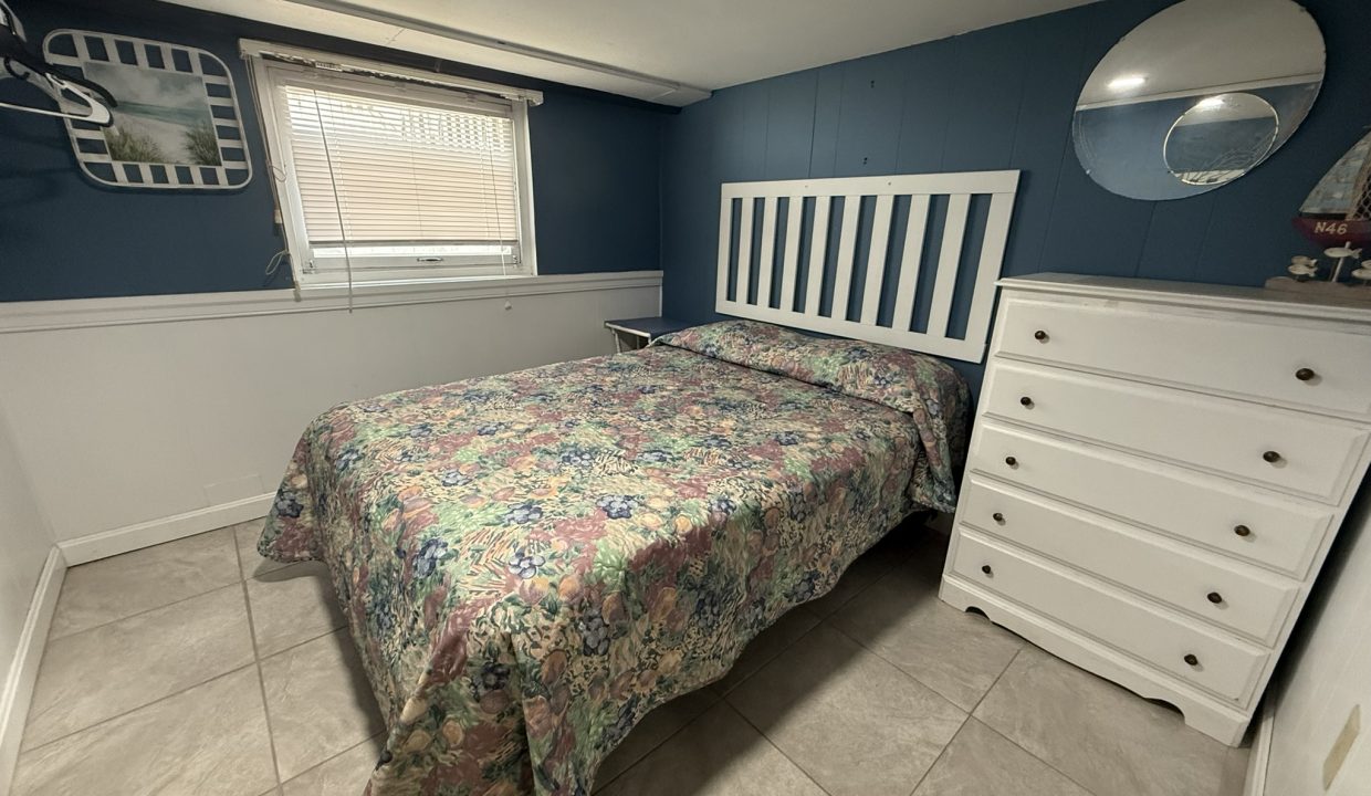 A small bedroom with a bed covered in a floral bedspread, a white headboard, a white dresser, a mirror, wall art, and a window with blinds.