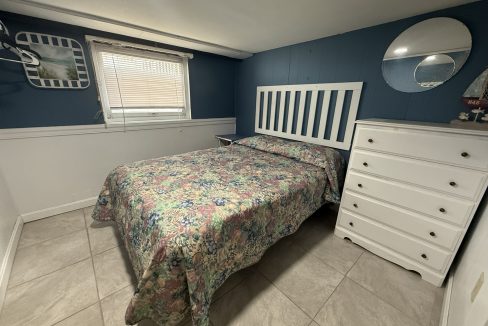 A small bedroom with a bed covered in a floral bedspread, a white headboard, a white dresser, a mirror, wall art, and a window with blinds.