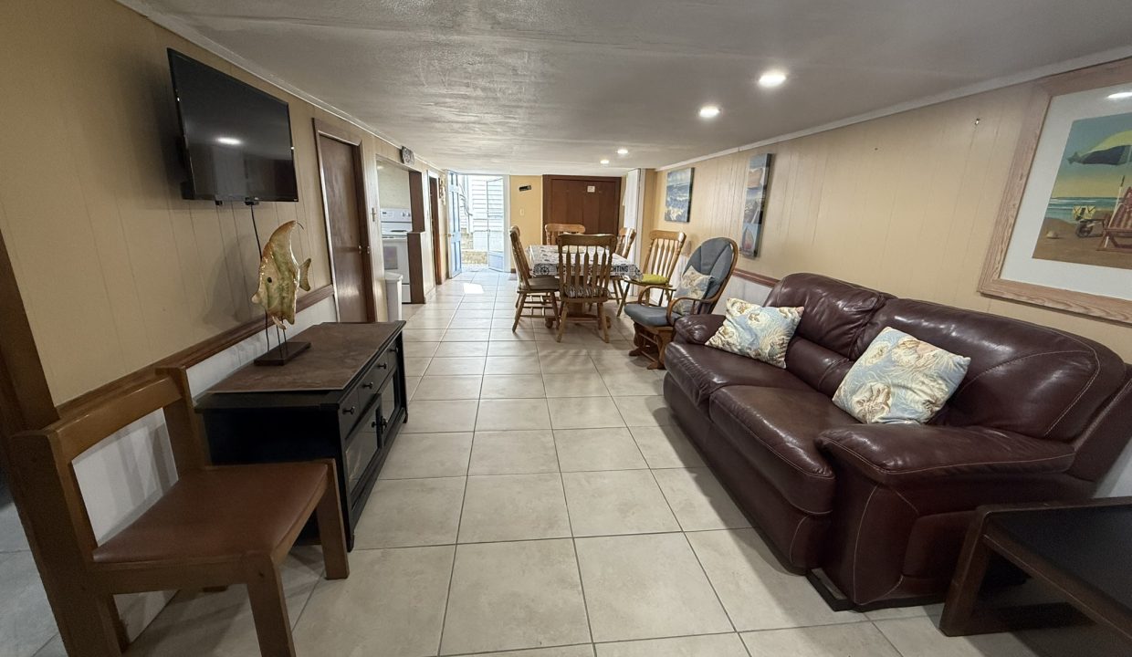 A long, tiled living room with a brown leather sofa, wooden chairs, a dining table, wall-mounted TV, and framed artwork on beige walls.