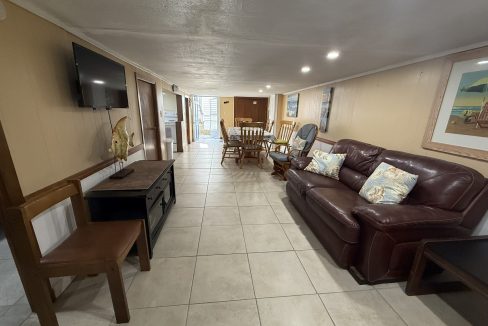 A long, tiled living room with a brown leather sofa, wooden chairs, a dining table, wall-mounted TV, and framed artwork on beige walls.