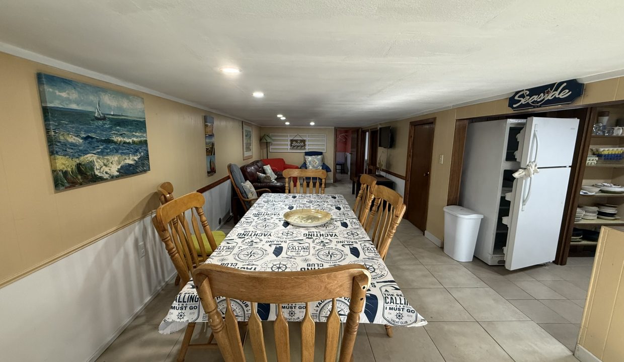 A dining area with a table and six wooden chairs, nautical-themed tablecloth, wall art, a white fridge, shelves, and a small living area in the background.