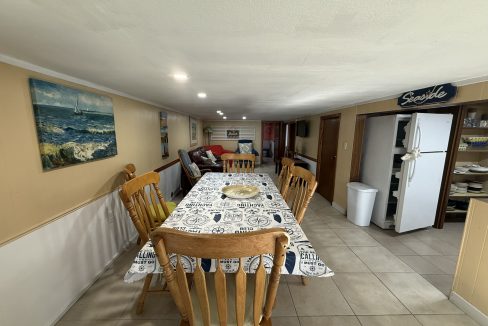A dining area with a table and six wooden chairs, nautical-themed tablecloth, wall art, a white fridge, shelves, and a small living area in the background.