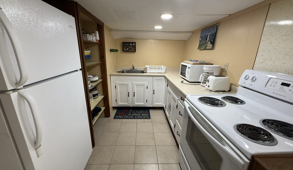 Small kitchen with white appliances, open shelving, a sink, countertop with microwave and toaster, and tile flooring; ceiling lights provide illumination.