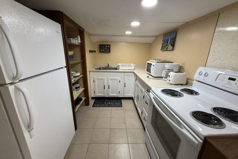 Small kitchen with white appliances, open shelving, a sink, countertop with microwave and toaster, and tile flooring; ceiling lights provide illumination.