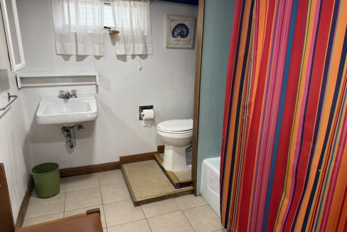 A small bathroom with a white sink, toilet on a raised platform, green trash bin, and a shower with a colorful striped curtain.