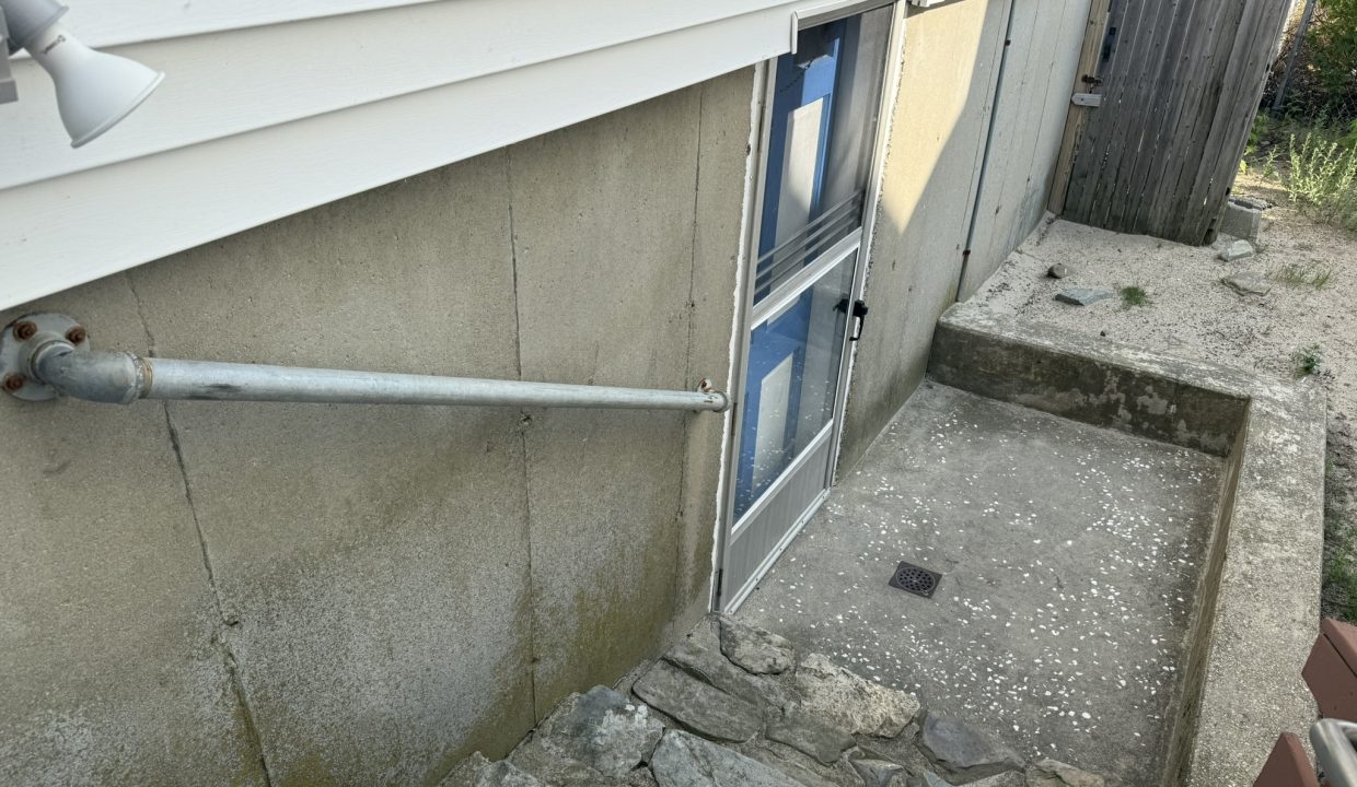A concrete basement entryway with rock steps, a metal drain cover, a pipe along the wall, and a closed door covered partly in blue tape.