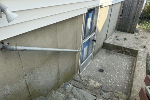 A concrete basement entryway with rock steps, a metal drain cover, a pipe along the wall, and a closed door covered partly in blue tape.