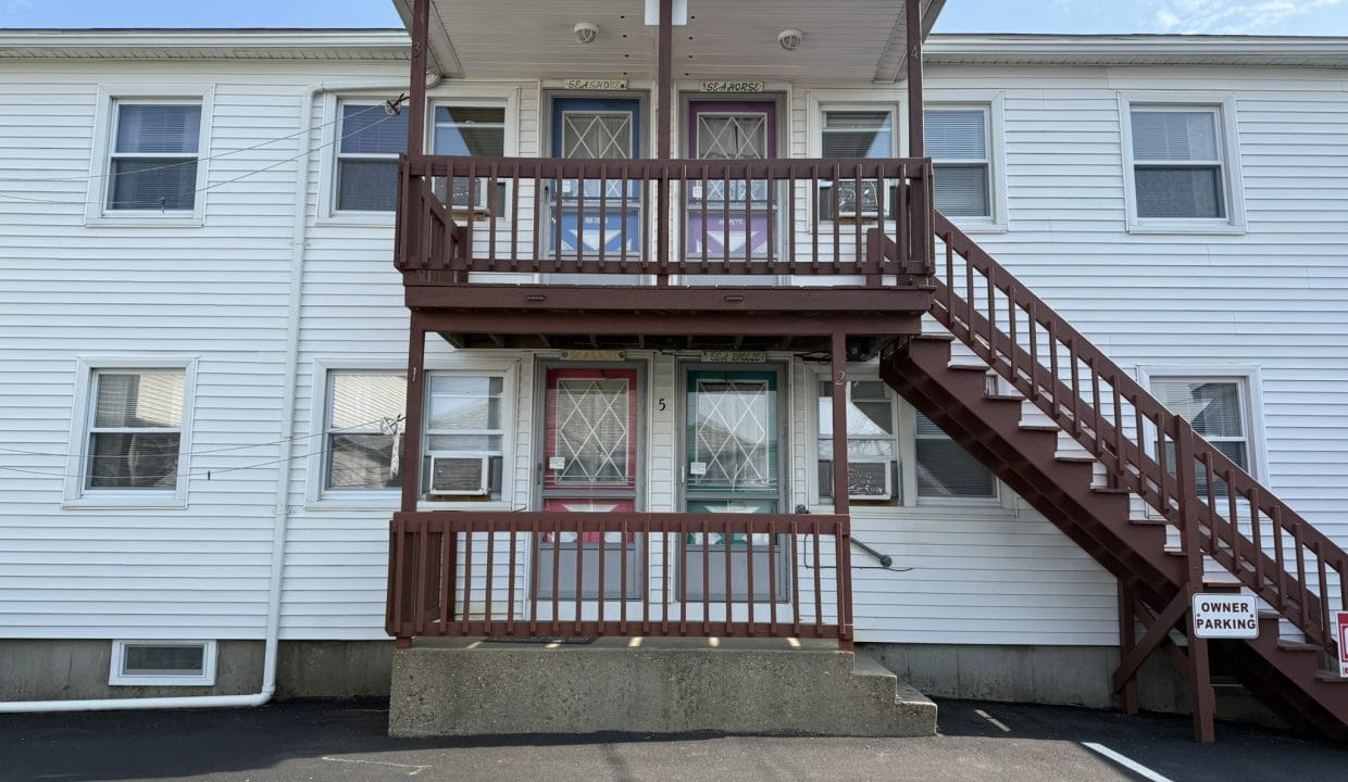 Two-story white apartment building with a central staircase, two doors on each level, and a sign marking 