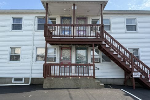 Two-story white apartment building with a central staircase, two doors on each level, and a sign marking 